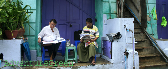 Two men reading news paper in front of a blue house" title="Two men reading news paper in front of a blue house